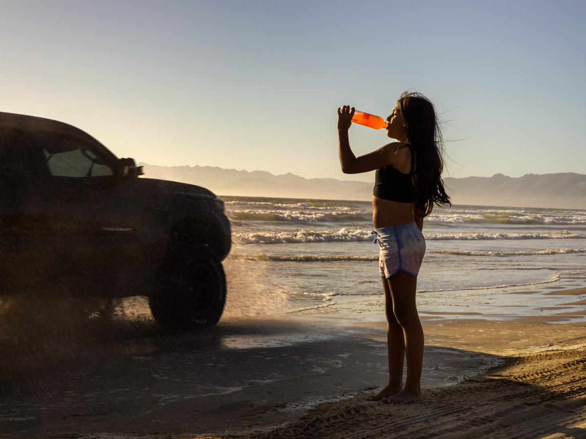girl drinking orange drink on beach with truck passing